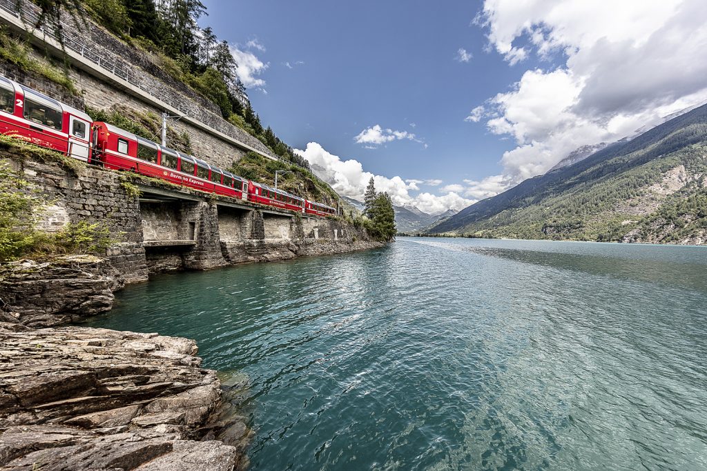 Bernina Express sightseeing train runs past scenic Lake Poschiavo in Switzerland.