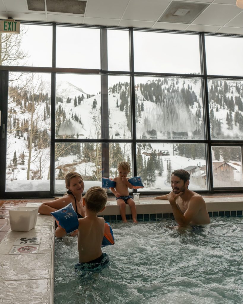 Family of four in hot tub with snowy capped mountain outside window.