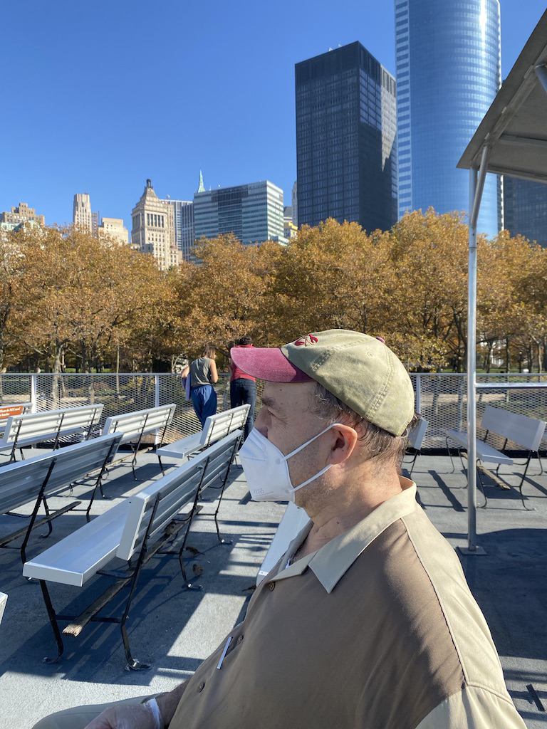Man in face mask sits on bench with New York City skyline in background.