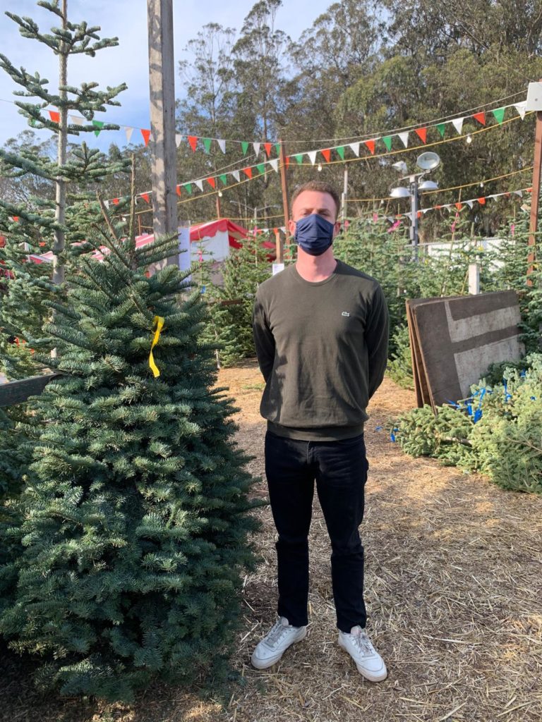 Man in face mask stands with selected Christmas Treet at u-pick farm.