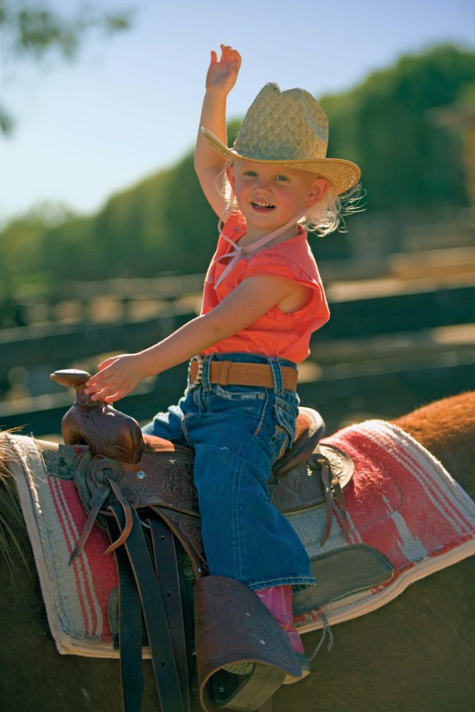 Young girl sits on top of a horse and waves like a bronco rider.