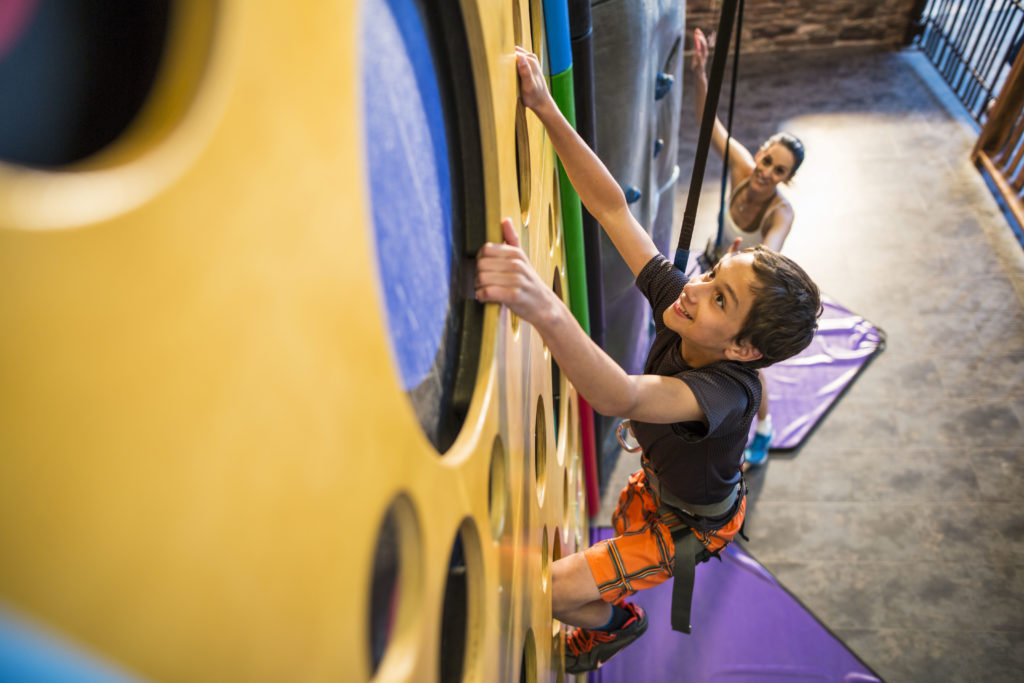 Boy climing an indoor rock climbing wall while woman watches him from the ground.