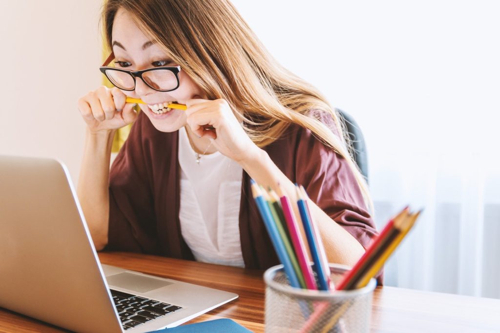 Asian woman biting pencil in front of laptop looks very annoyed.