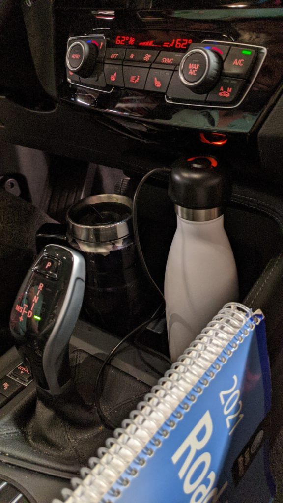 Car dashboard with travel coffee mug and water bottle plugged into charger.