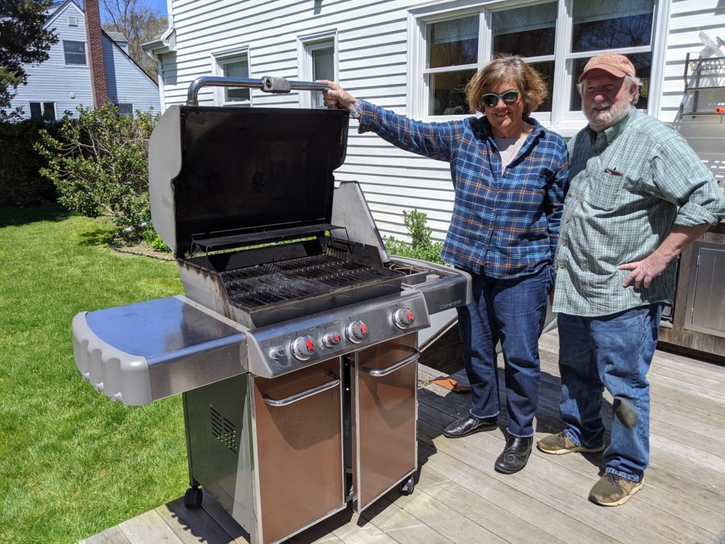 Couple standing next to a large Weber propane barbecue with burners.