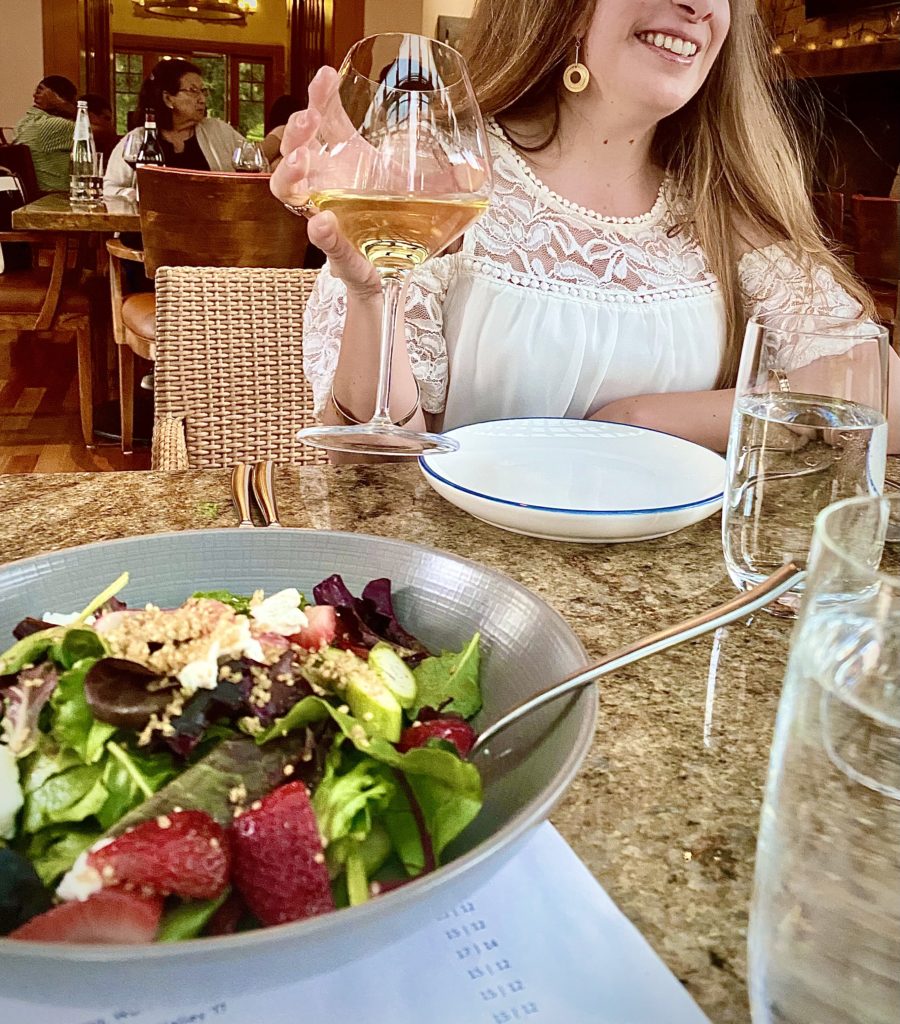 Woman drinking glass of wine at a dinner table with salad in the foreground.