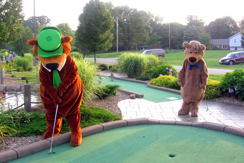 People in Yogi Bear and Boo-Boo Bear suits play miniature golf together at a Jellystone Park.