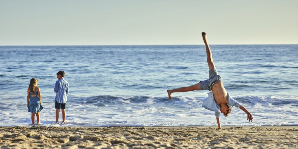 Kids stand at water's edge while a man does cartwheels on beach.