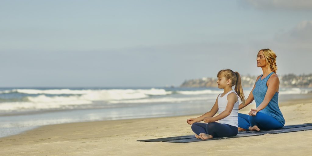 Girl and mom meditate in seated pose on the beach.
