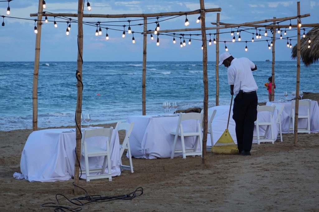 Man sweeping the beach as tables are set on the sand for a romantic dinner.