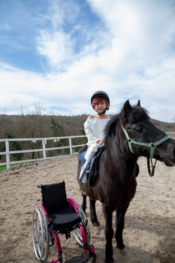 Girl on black pony in corral with wheelchair next to her