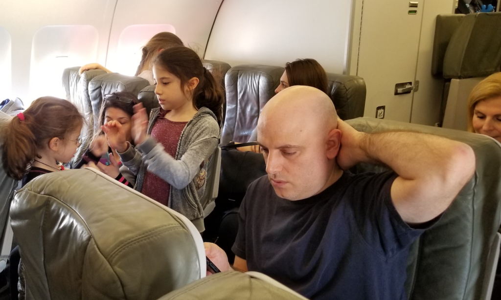 Busy dad is rading and his girls playing in the aisle of an airplane.
