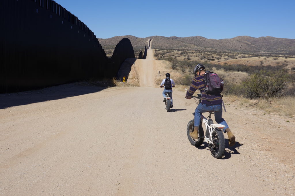 Two men on e-bikes ride along the border wall between Sasabe, Arizona and Sasabe Pueblo, Mexico.