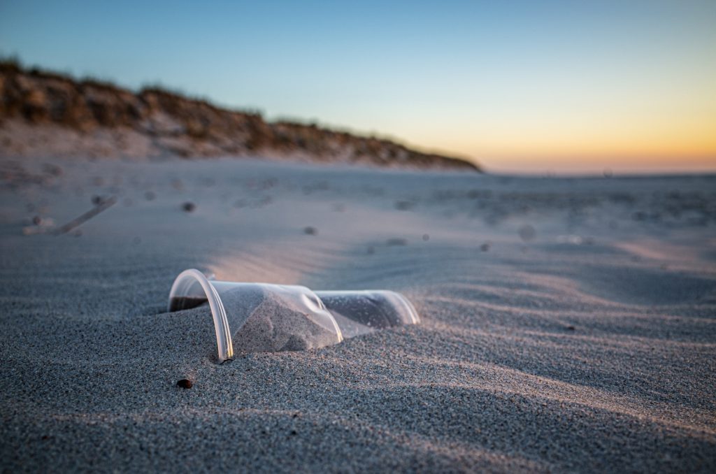 Plastic drinking cup is buried in the sand at the beach. By hamsterfreund via pixabay.