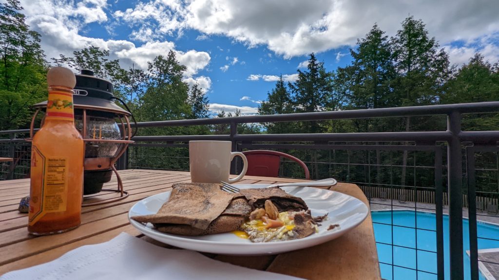 Plate of breakfast crepes and hot sauce on a picnic table, pool in background.