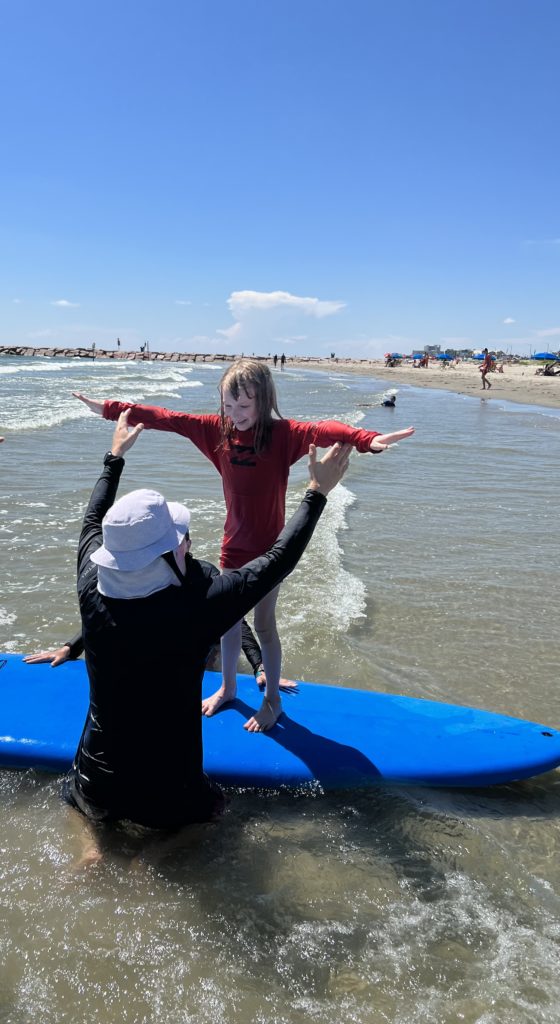 Mom and teacher shows girl how to stand up on a surf board in shallow surf.