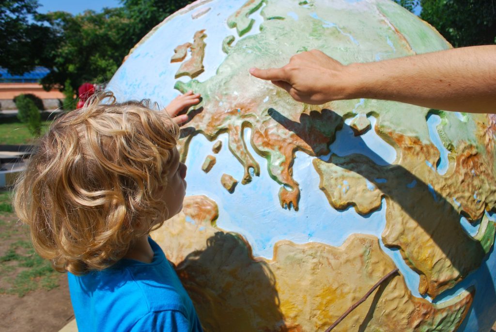 Adult points out a country in Europe on a huge outdoors globe to a young blond child.