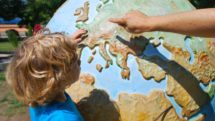 Adult points out a country in Europe on a huge outdoors globe to a young blond child.