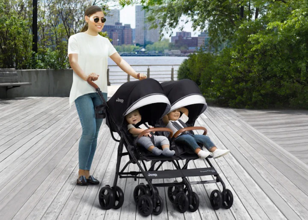 A Jeep Powerglyde Plus Stroller with twins in it and a mom standing by.