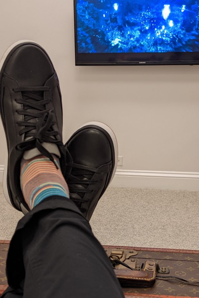 A person with legs crossed on top of luggage watches TV screen.