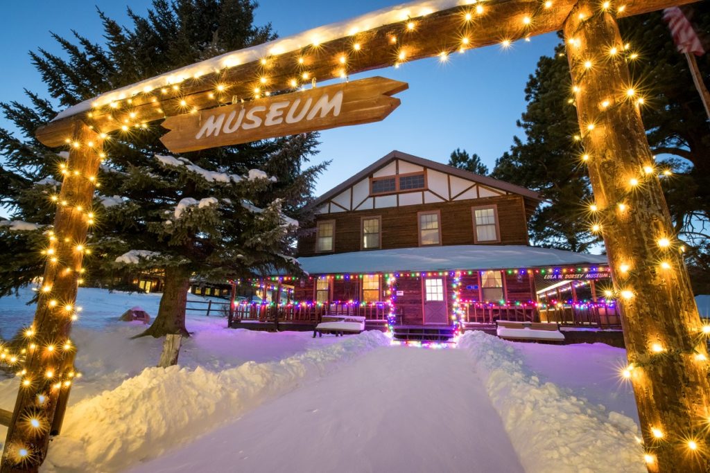 Log cabin covered in Christmas lights, surrounded by snow, with an entry archway and museum sign.