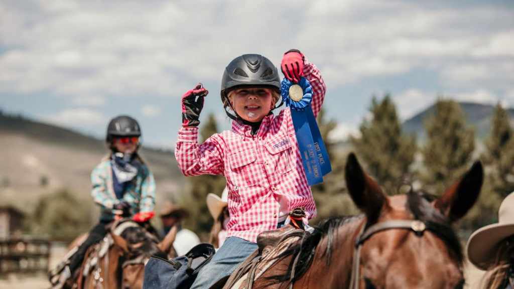 Child on horseback holds blue ribbon for riding skills.