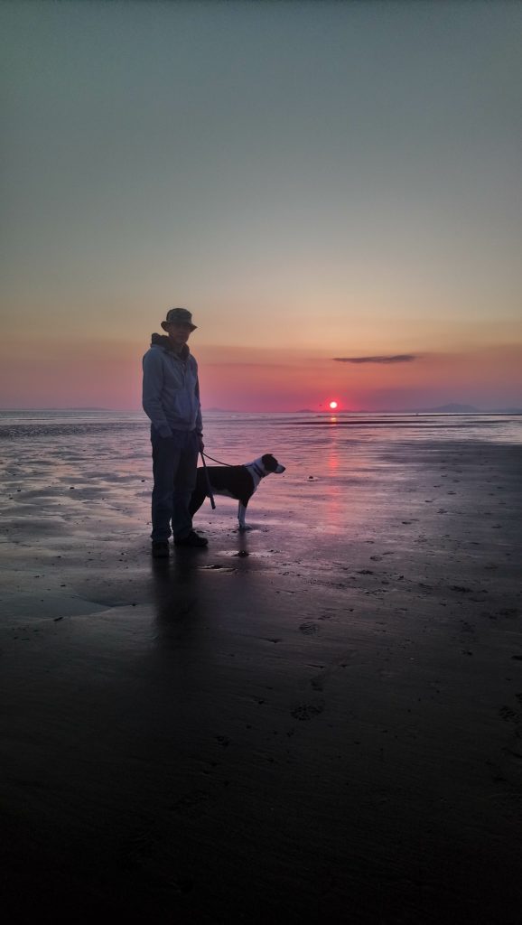 Man enjoyng the sunset at the beach with a dog on leash.