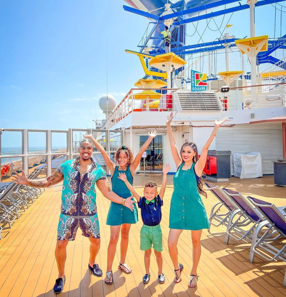 Family with couple and two children in summer outfits are posing with hands in the air on Carnival cruise ship deck.