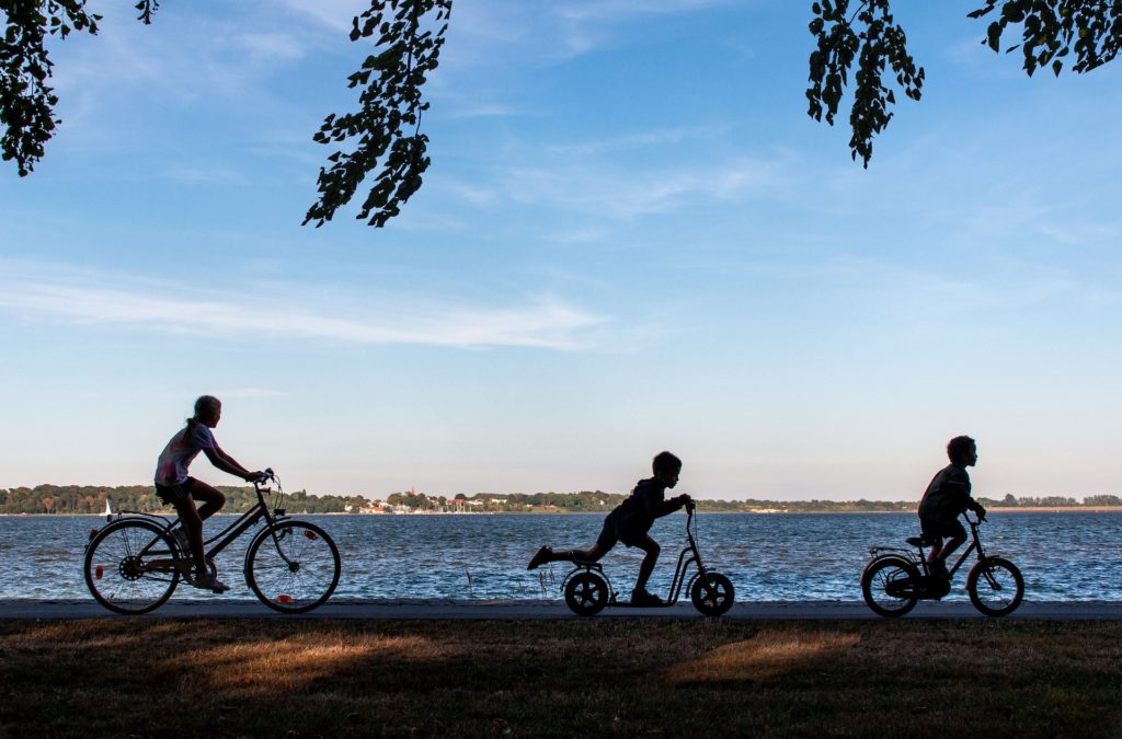 Adult on a bike and two kids, one on a scooter and one on a bike, all in silhouette, ride along the waterfront of a park.