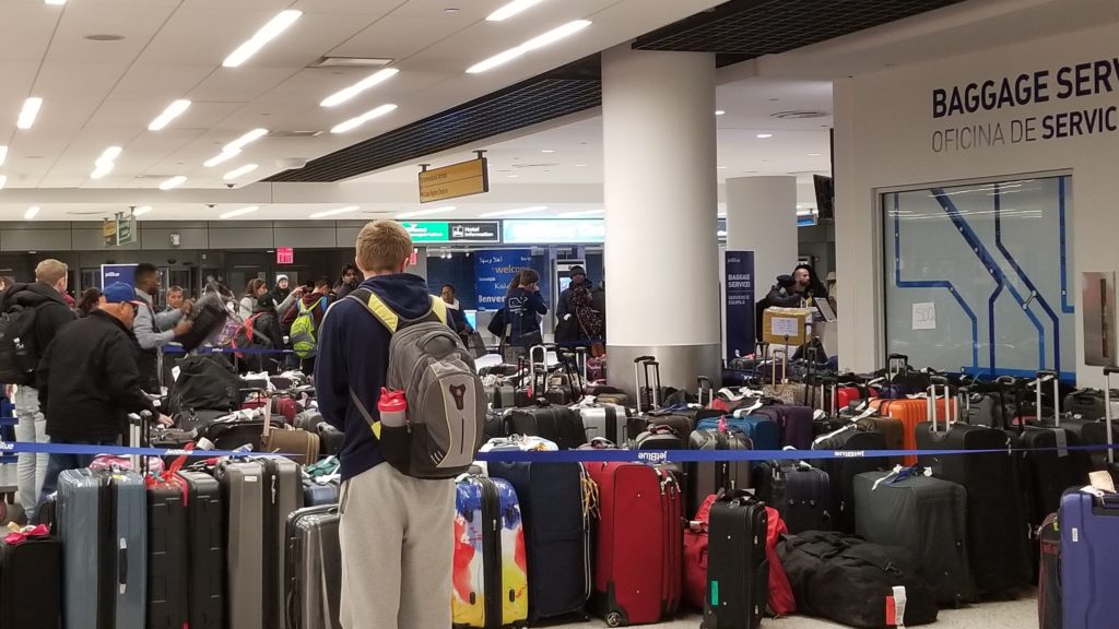 Man with backpack searches for his luggage among a large collection of suitecase in a crowded airport baggage claim.