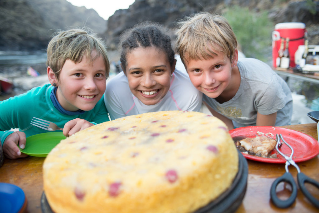 Three kids outdoors smile while looking at a freshly baked cake.
