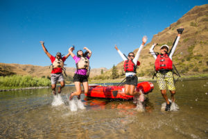 Four people jump up and down in the water next to a kayak.