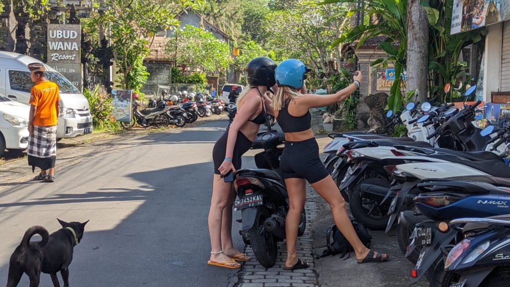 Two female tourists post for selfies with their moped and helmets on a street in Ubud, Bali.
