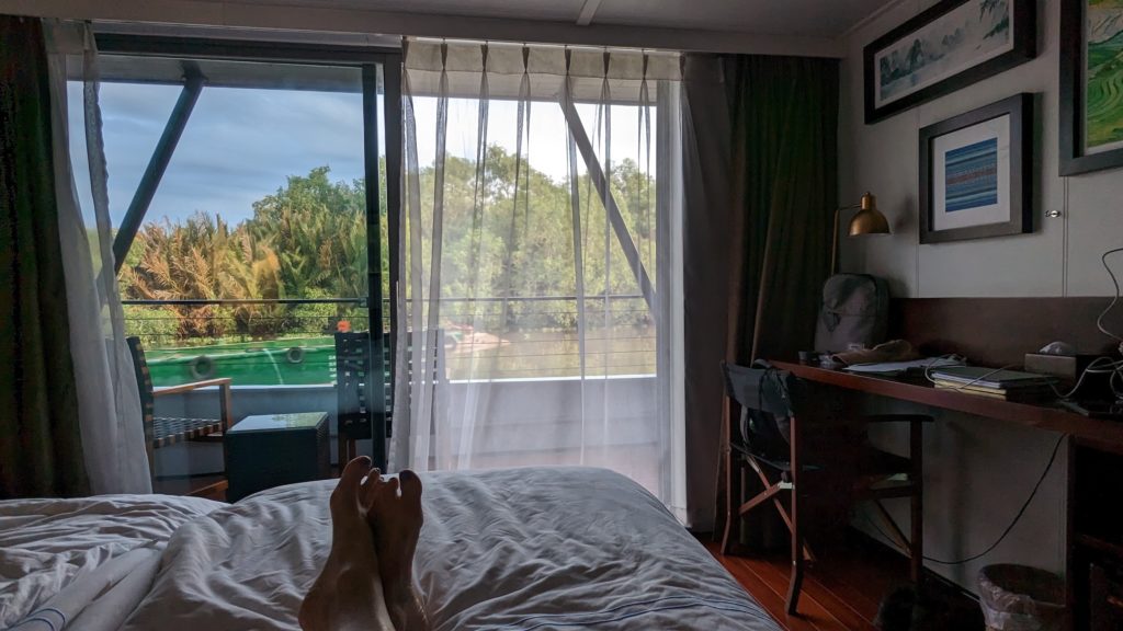 Person's feet on bed in front of view of Mekong River from a river cruise ship cabin.