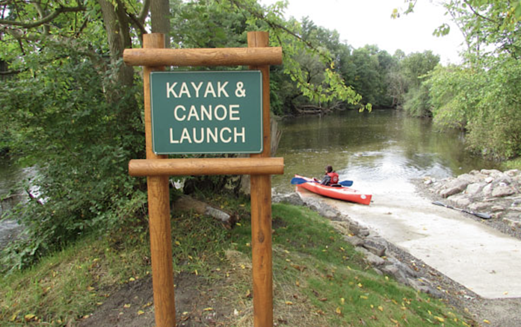 Kayak and canoe launch by the side of a river with kayaker in the background.