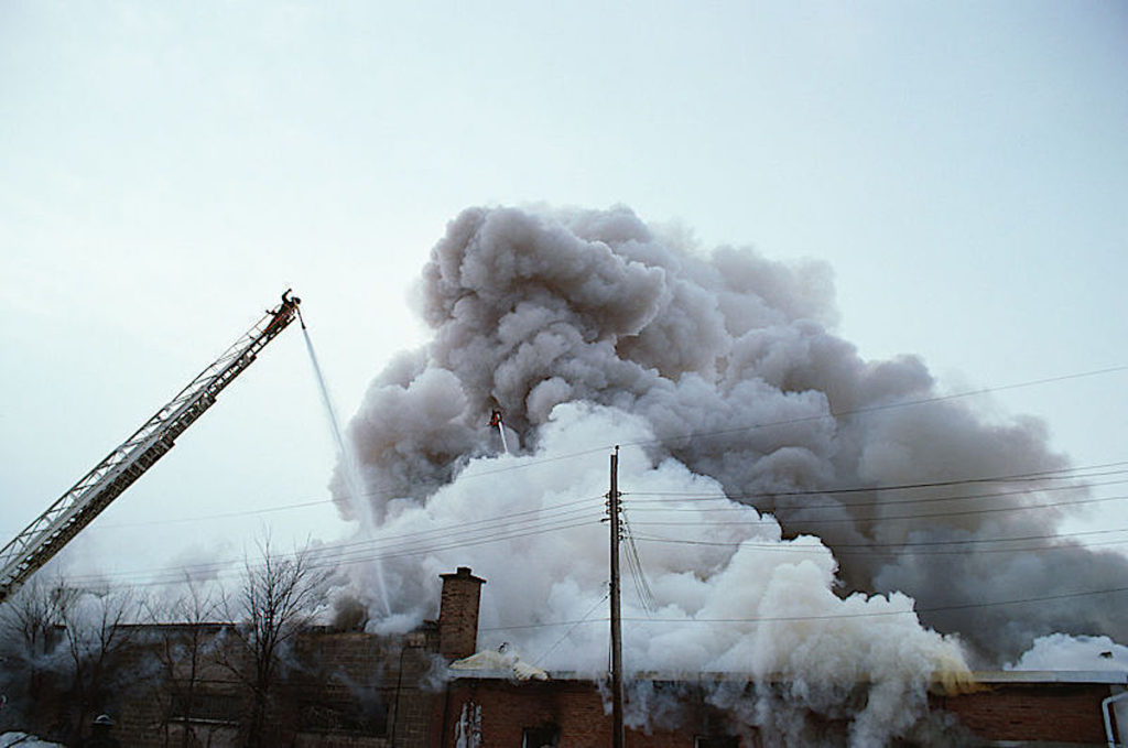 Factory on fire with firefighter spraying water and billowing smoke.