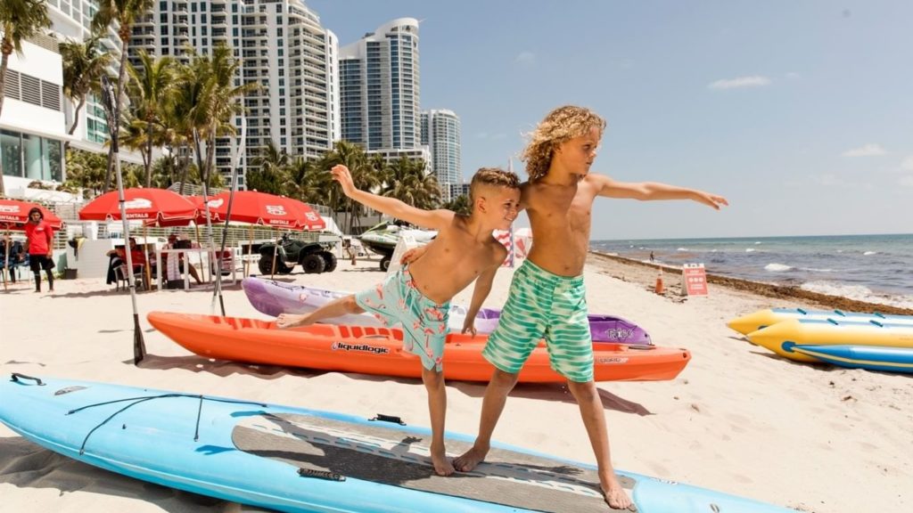Two boys playing on top of a paddleboard at the Diplomat Hotel on a Florida beach.