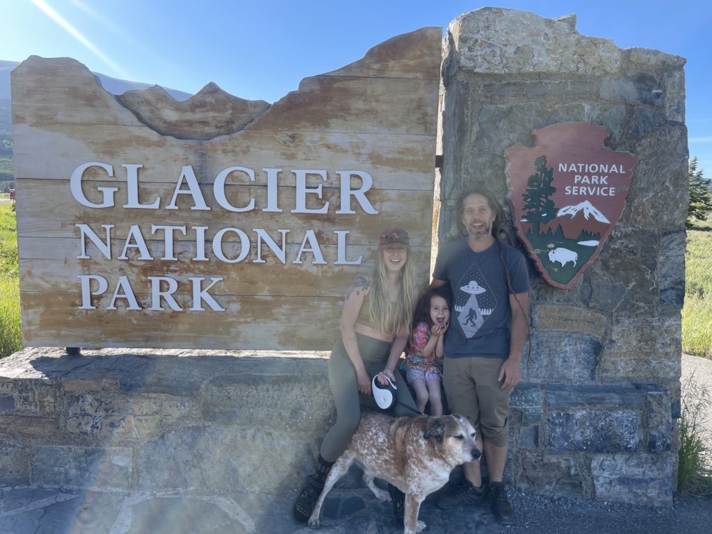 Family and dog pose in front of sign for Glacier National Park, Montana.