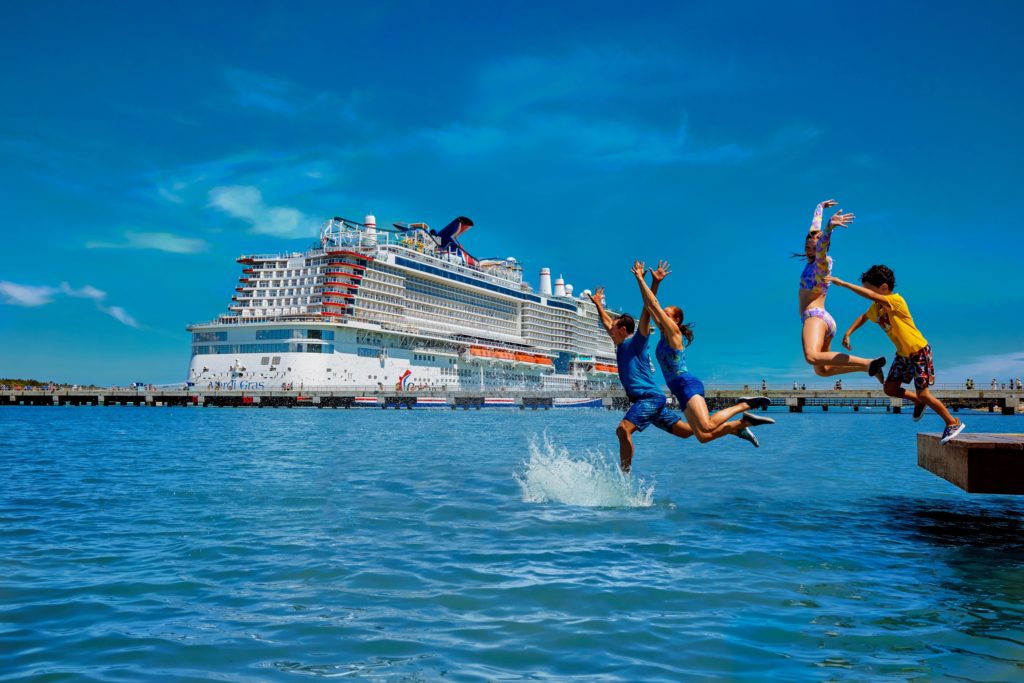 Kids jump into the water with a Carnival cruise ship in the background at sea.