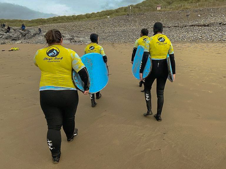 Four people in wet suits walk to the surf of Sligo carrying surfboards.