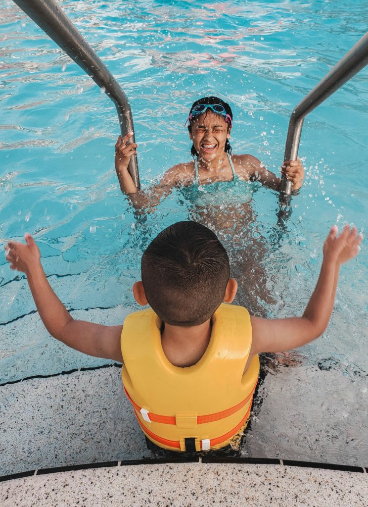 Boy in life jacket sits at edge of pool playing with woman in the water.