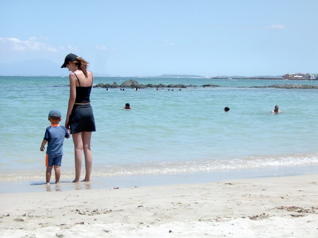 Mother and son at water's edge in Capetown, wearing sun protection clothes and hats.