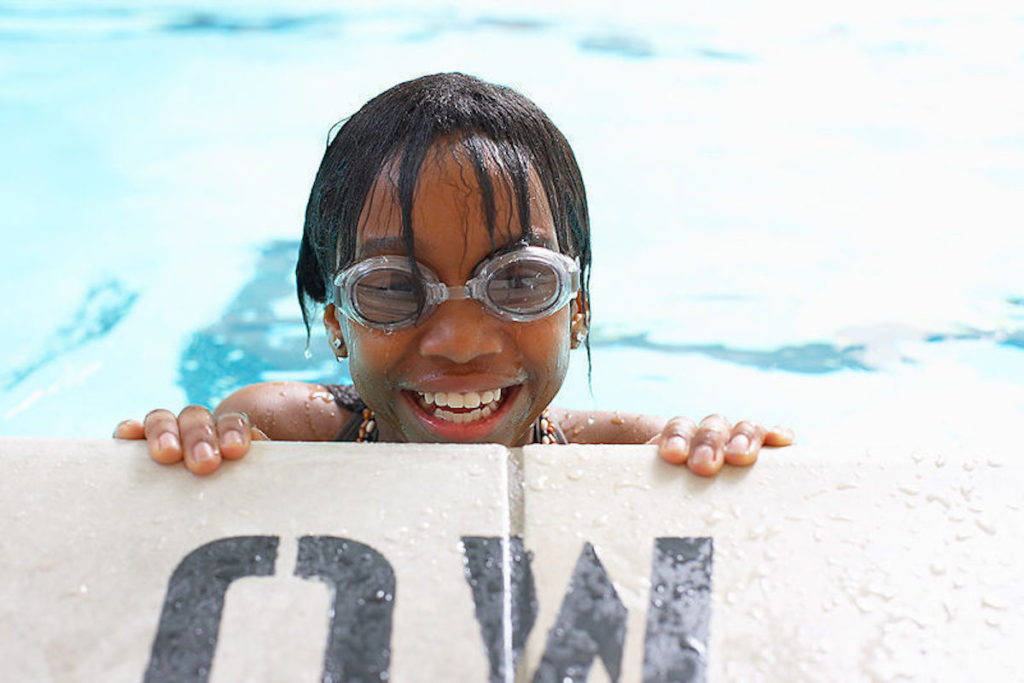 Teen girl wearing swim goggles smiles at camera from the edge of the pool