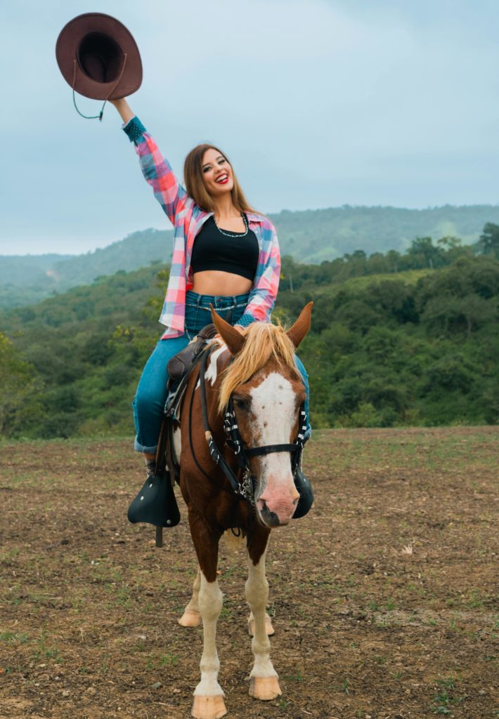 Woman waving hat enjoys horseback on a mountain trail. Photo c. Daniel Gonzalez for pexels.