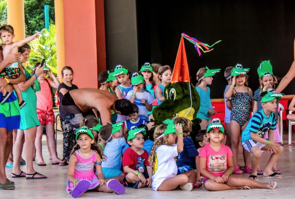 The alligator mascot for the Vivitos Kids Club poses with a group of kids in alligator hats outside the kids club at a Viva by Wyndham resort. Photo c. Viva Wyndham