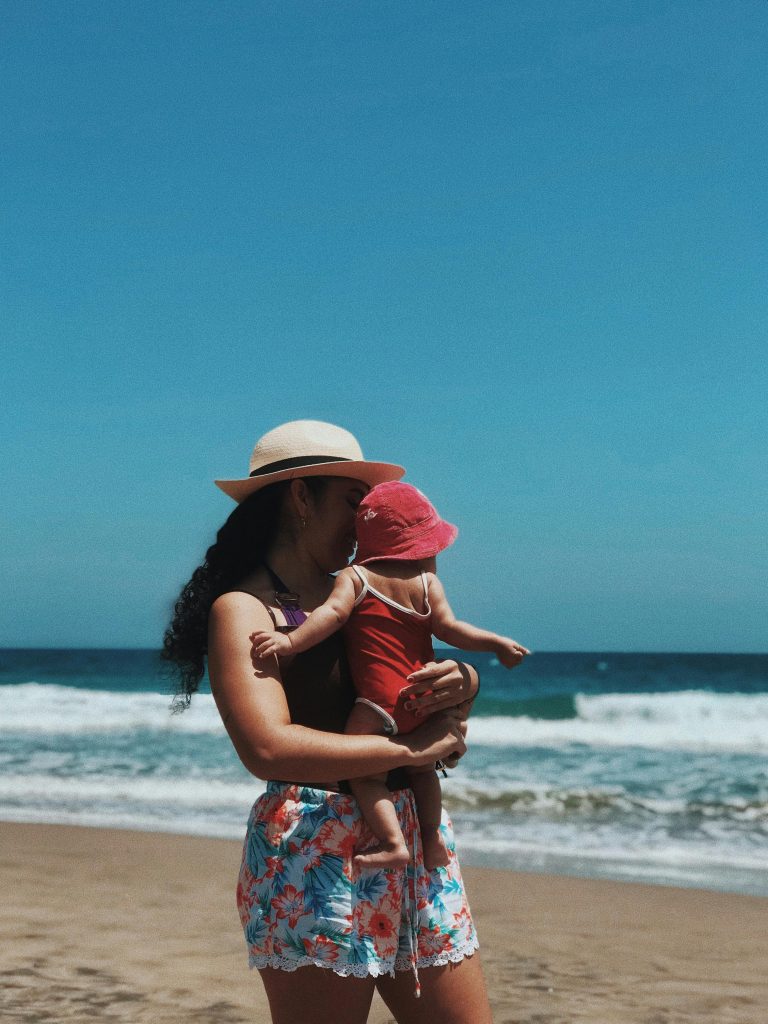 Mom and straw hat holds baby in beach hat while walking on the beach. Photo c. Ciindy Veliz Army for pexels.