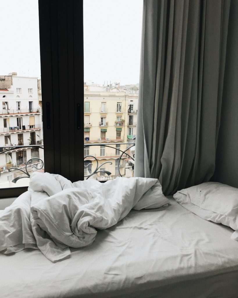 An unmade bed with white covers by a window with a view of the Ciutat Vela neighborhood of Barcelona. Photo by Lina Kivaka for pexels.