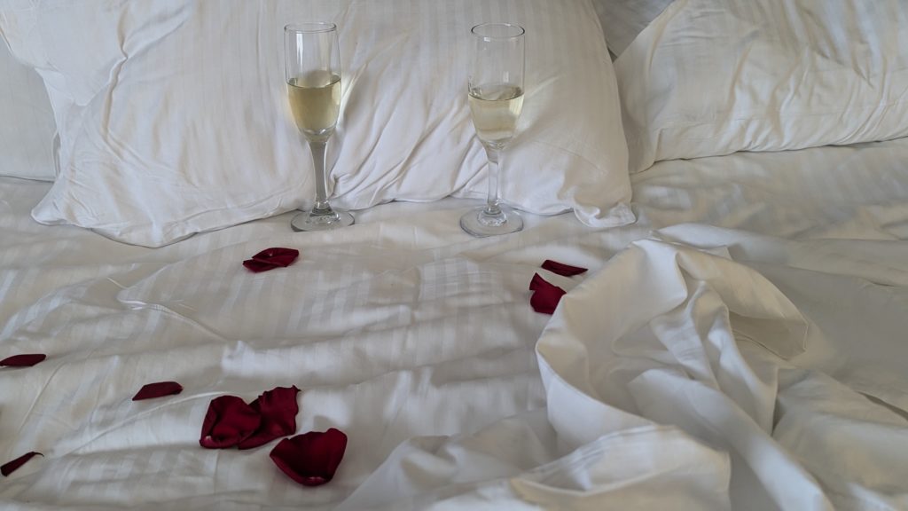 Two glasses of champagne and messy bed scattered with rose petals.