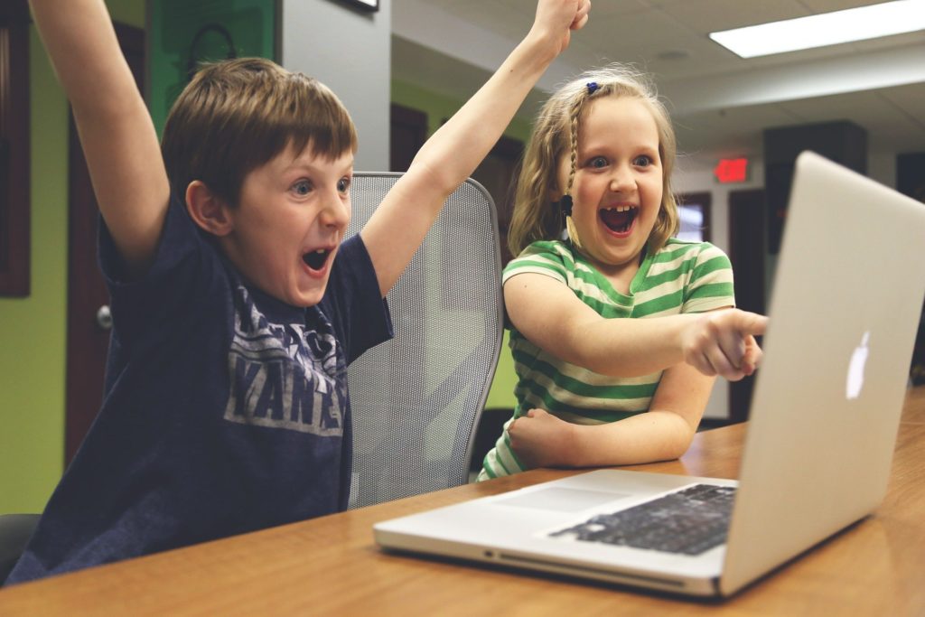 Two kids playing on a laptop are cheering at what they see.