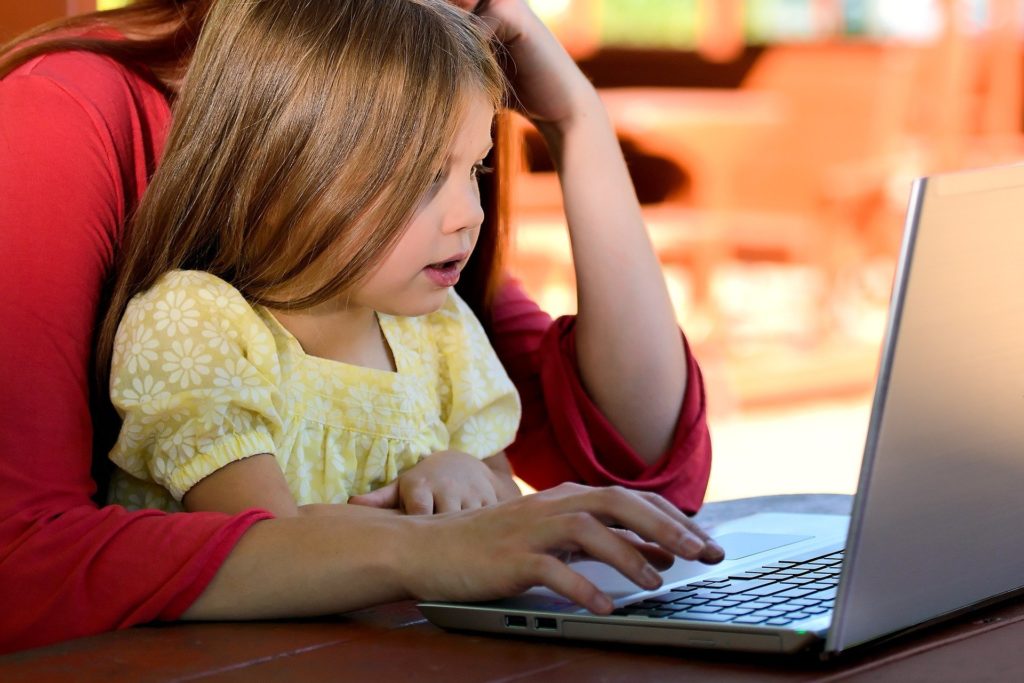 Mom sits at laptop computer with young girl on her lap.